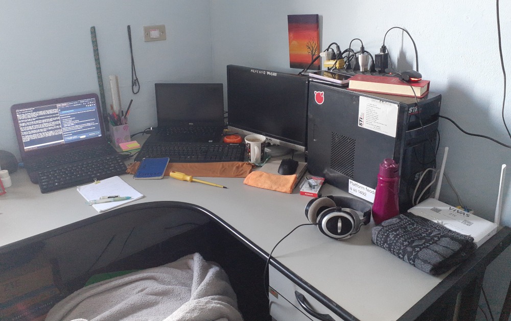 An L-shaped desk with two laptops, an external monitor, a router and a third headless computer in a tower case with several power cables connected to a power strip on top of it. Next to the power strip are two cellphones, a long red box, and a charging case for Bluetooth headphones with a red LED on. The tower case has three stickers on it: one with the machine specifications, one the FreeBSD logo, and one reading “platform feodalism (sic) is so 1492”. Scattered around the machines are some office supplies, medicine containers, a screwdriver, a notepad, a small notebook, an NVMe SSD card on top of its packaging, a mug, a water bottle and a scarf. Hanging on the wall are a small painting of a dead tree before the twilight, prayer beads, a sunflower-pattern keychain and a calendar. Between the desk and the camera, a green plastic chair has a pillow and blanket on top of it. A few wires and cardboard boxes are visible under the desk.