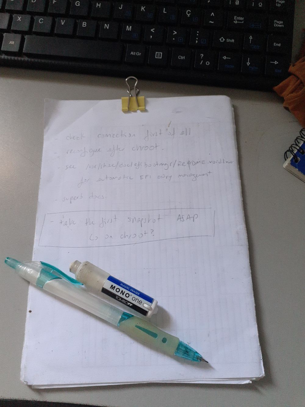 A block of paper with some notes scribbled: “check connection first of all”, “reconfigure after chroot”, “see /usr/share/doc/efibootmgr/README.voidlinux for automatic EFI entry management”, “superb docs”, “take the first snapshot ASAP”. An arrow points from the last note to “on chroot?” Visible above the block of paper is a keyboard. To the right, the tip of a notebook and a piece of brown cloth are visible. On top of the block, there is a mechanical pencil and a Tombow MONO One plastic eraser.